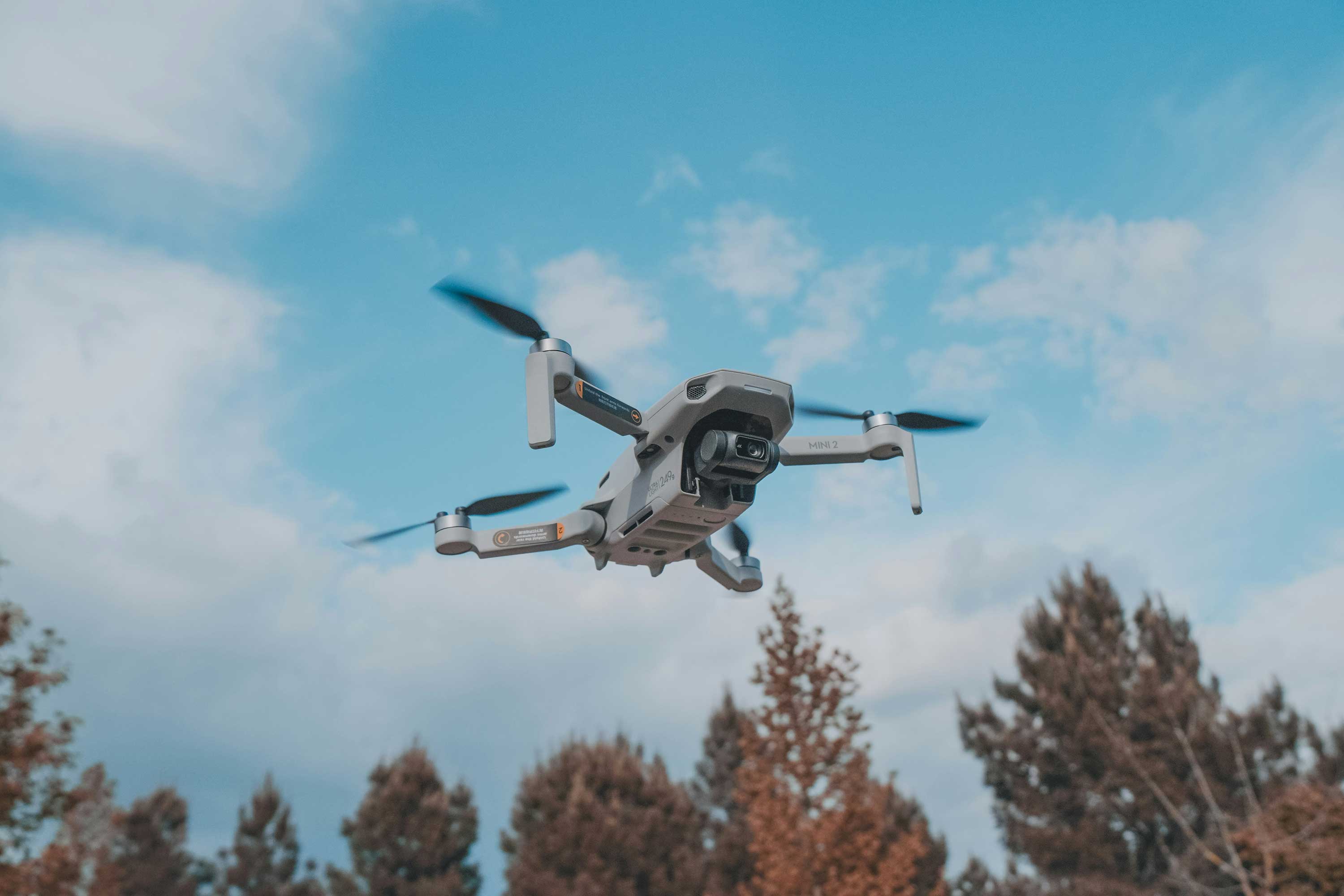 A DJI Mini 2 mid-flight with trees at the bottom and mostly blue sky behind